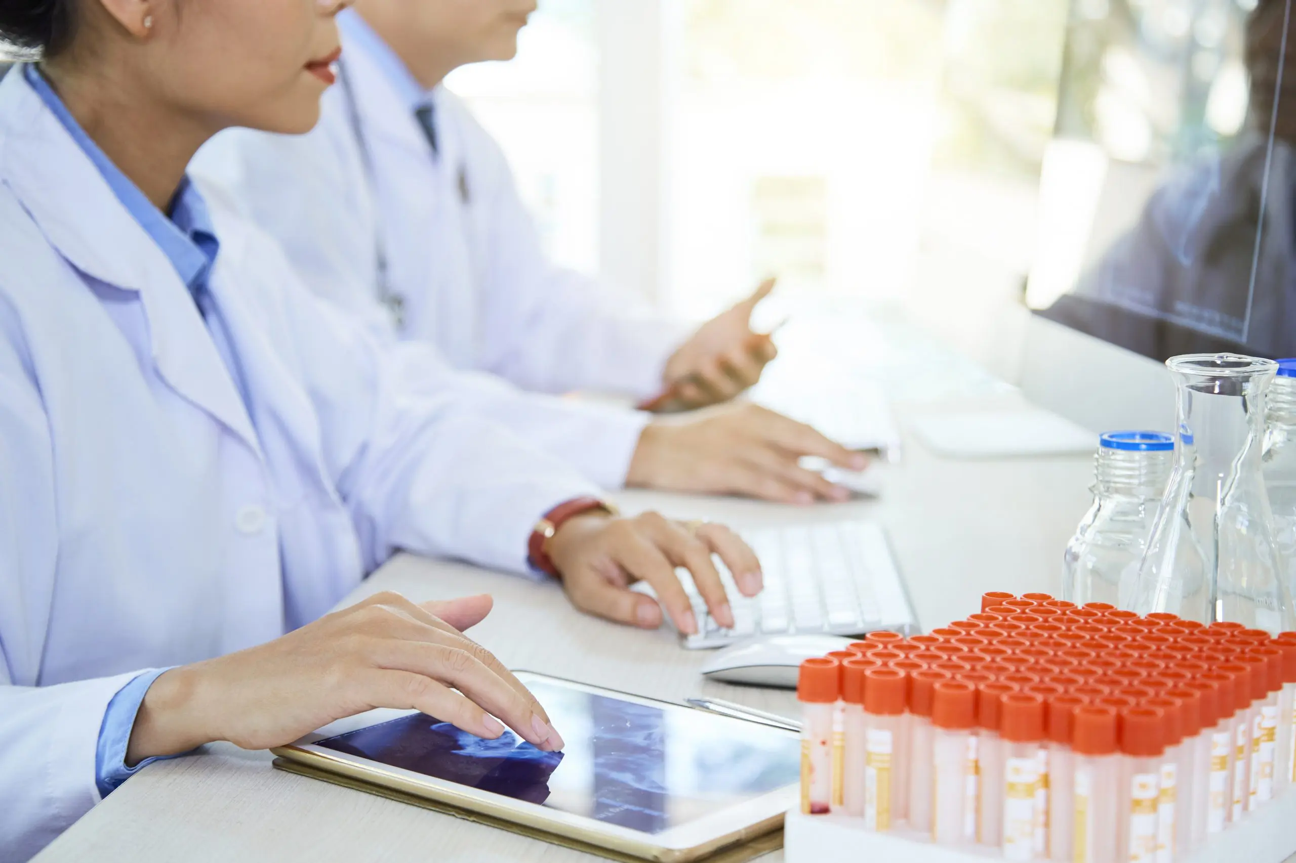 Close-up of female doctor sitting at the table with test tubes and using digital tablet with man working in the background at laboratory