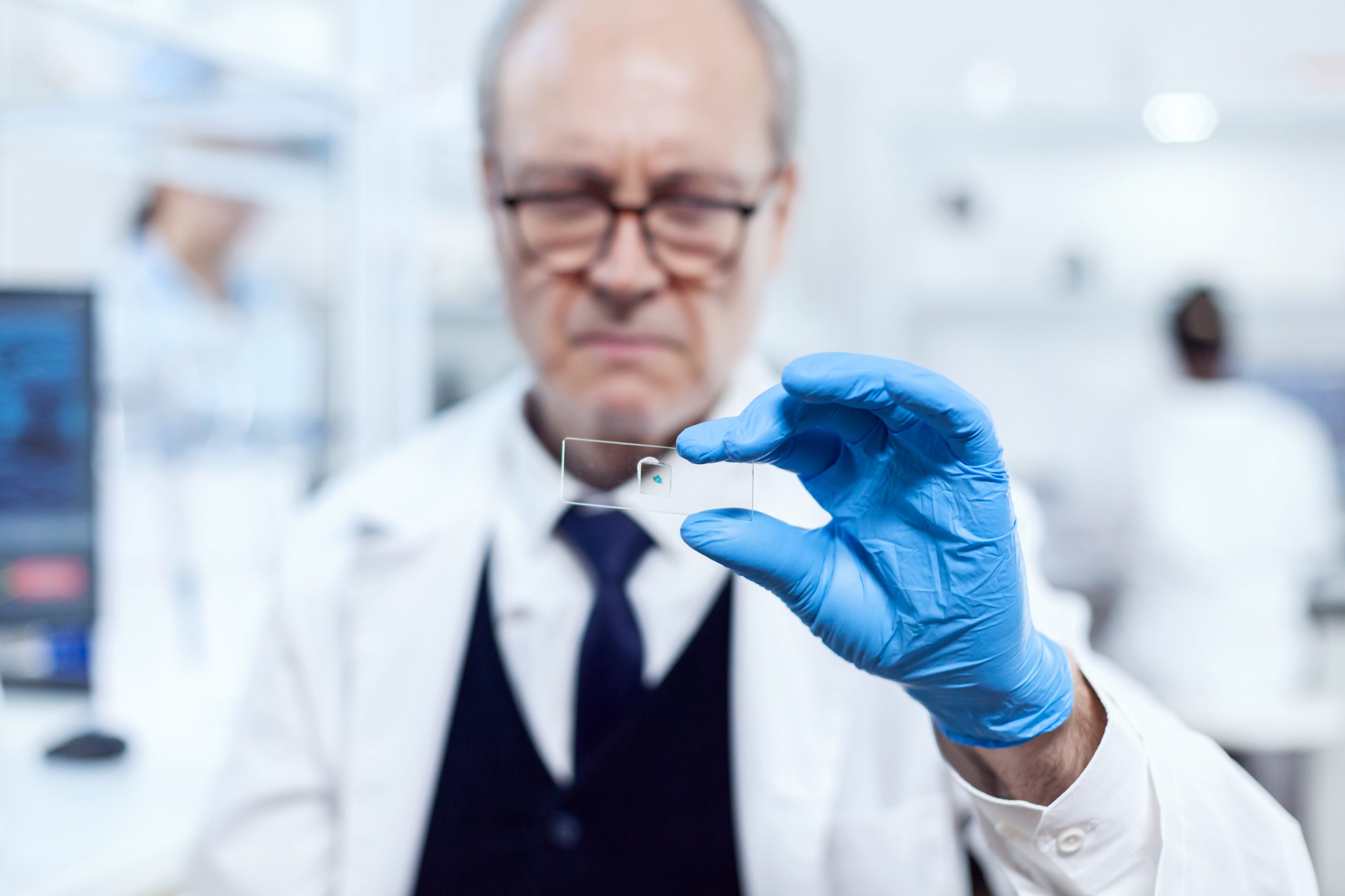 Close up of medicine sample on glass slide while scientist is looking at it in laboratory. Senior researcher in sterile lab looking on microscope slide wearing lab coat.